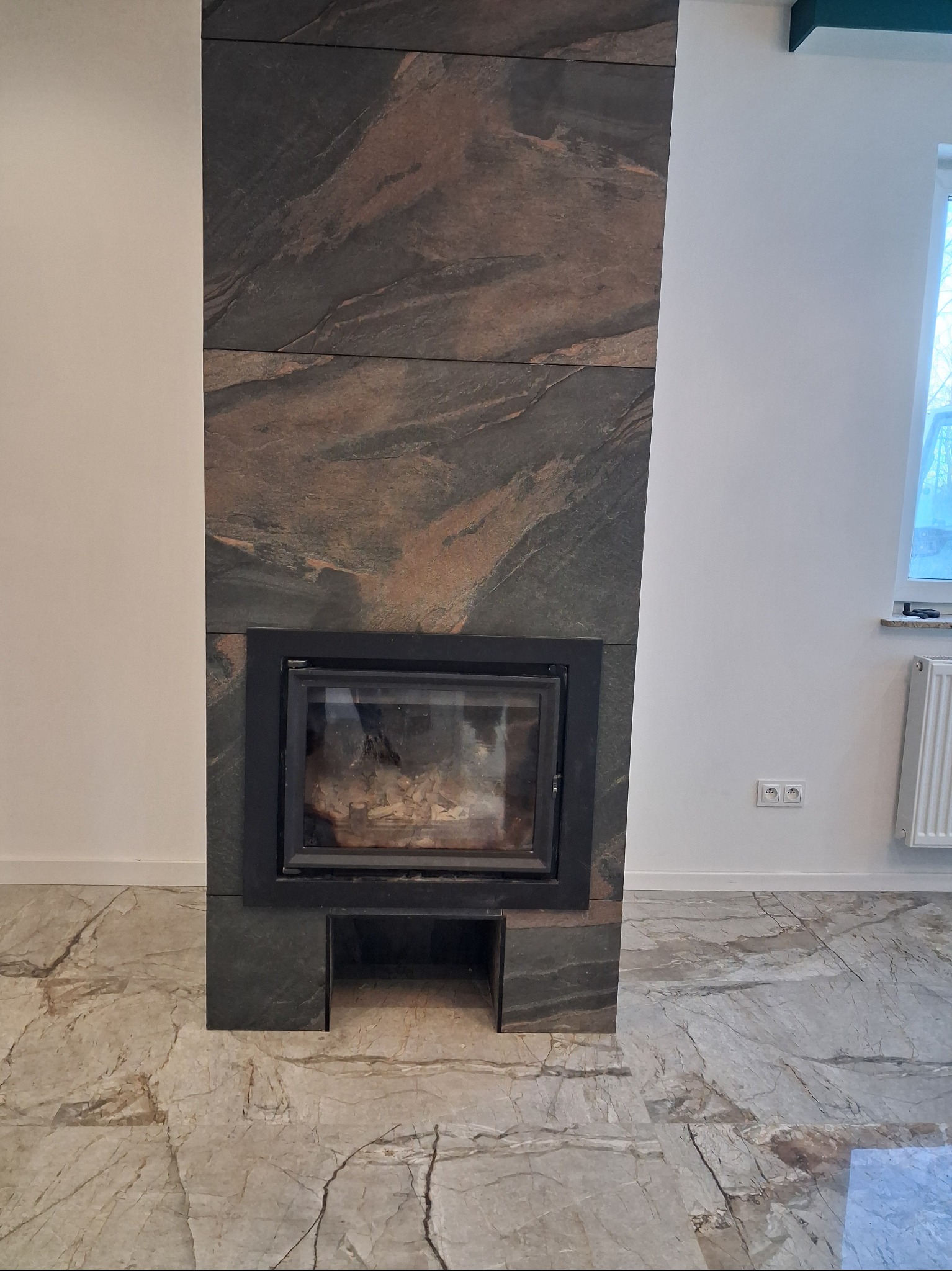 Nowoczesny kominek obłożony płytami z kamienia naturalnego o wyraźnym wzorze, dopasowany do marmurowej podłogi. Minimalistyczny design w jasnym wnętrzu.