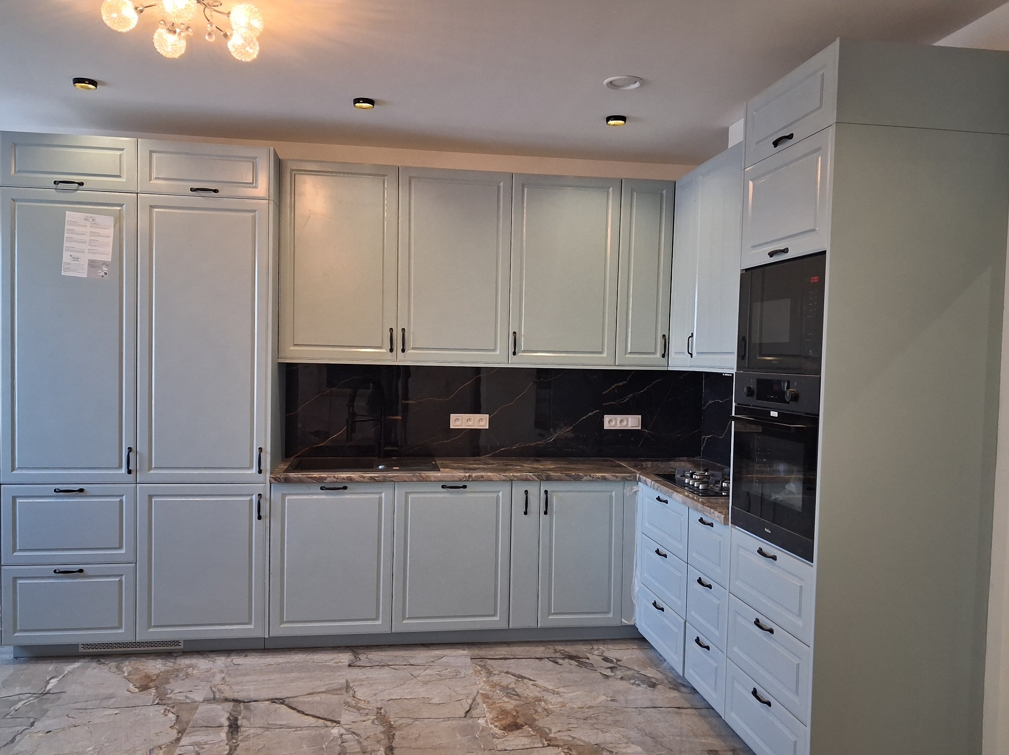 Jasna, pastelowa zabudowa kuchenna z czarnym blatem i marmurową podłogą. Uwagę zwracają frezowane fronty szafek i minimalistyczne, czarne uchwyty.