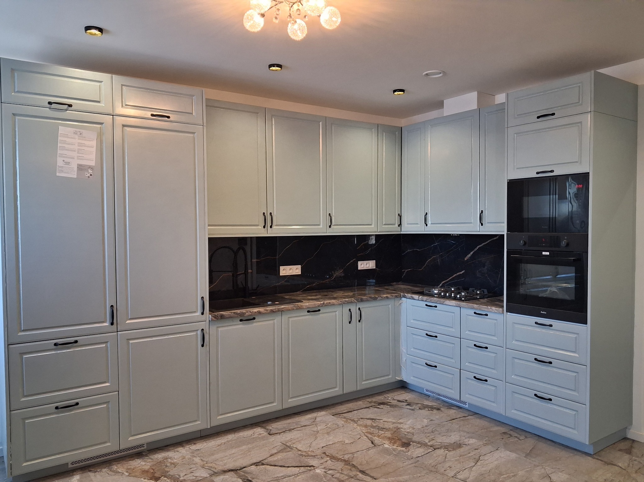 Kompletna, jasnoniebieska zabudowa kuchenna z czarnym blatem i sprzętami AGD. Klasyczny styl, marmurowa podłoga, oświetlenie sufitowe. Widok na całą kuchnię.