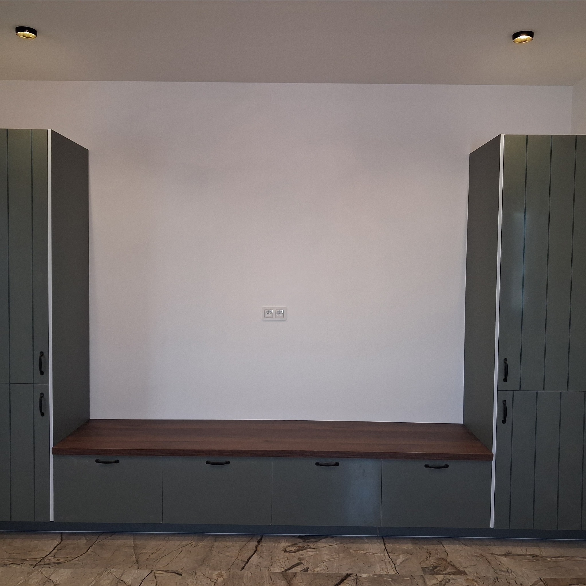 Zabudowa meblowa wnęki w odcieniach szarości z drewnianym blatem, widoczne gniazdka elektryczne na ścianie. Podłoga z wzorem imitującym marmur.