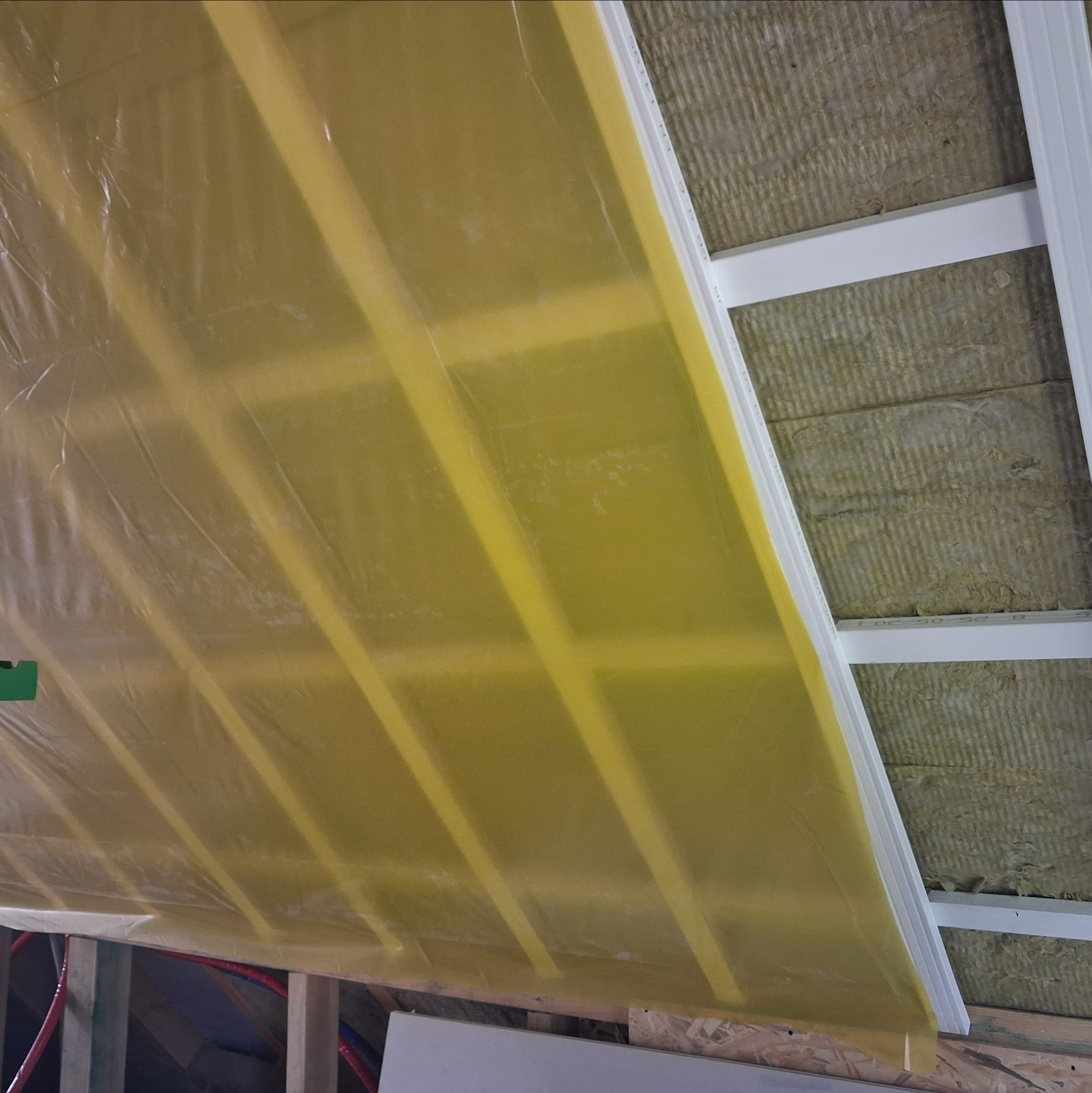 Ocieplenie poddasza wełną mineralną, zabezpieczone folią paroizolacyjną. Widoczne drewniane elementy konstrukcyjne i białe profile. Ujęcie z dołu, ukośne.