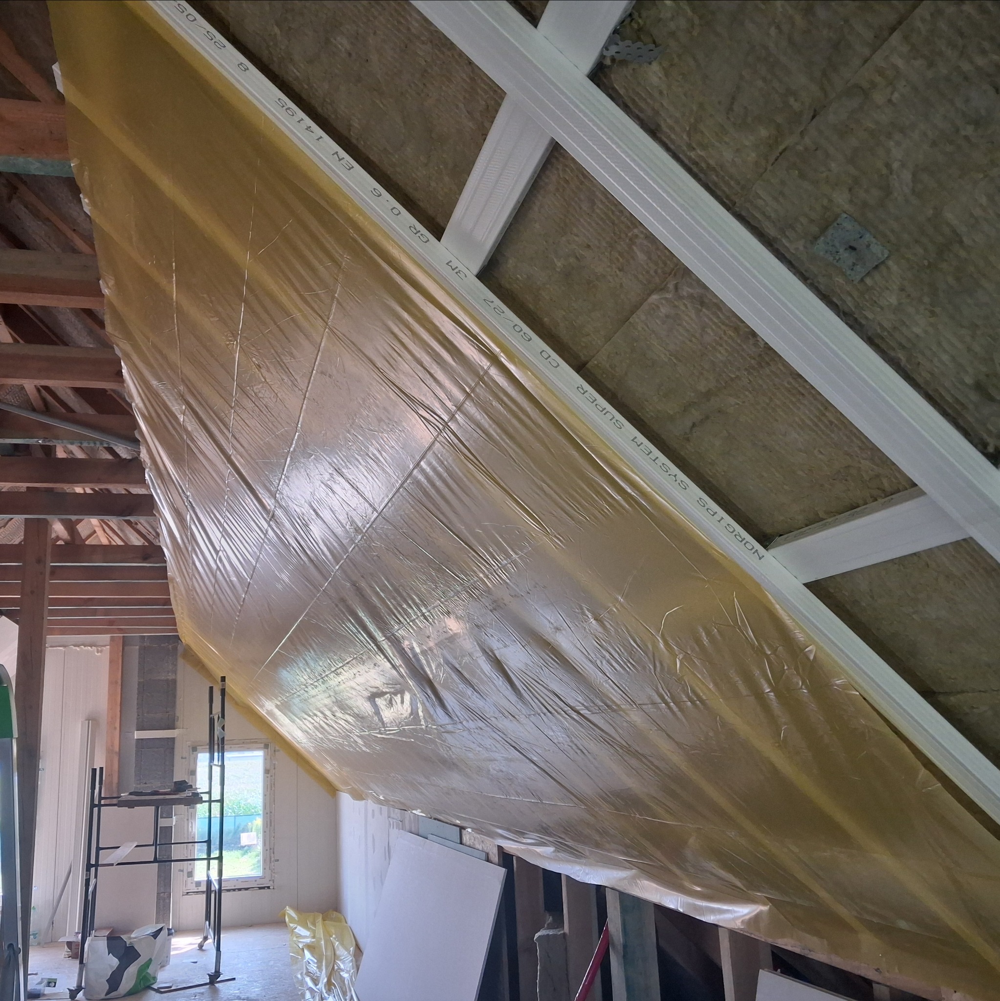 Ocieplenie poddasza z widoczną folią paroizolacyjną i wełną mineralną. Elementy konstrukcji drewnianej i metalowe profile sufitowe. Widoczne rusztowanie w tle.