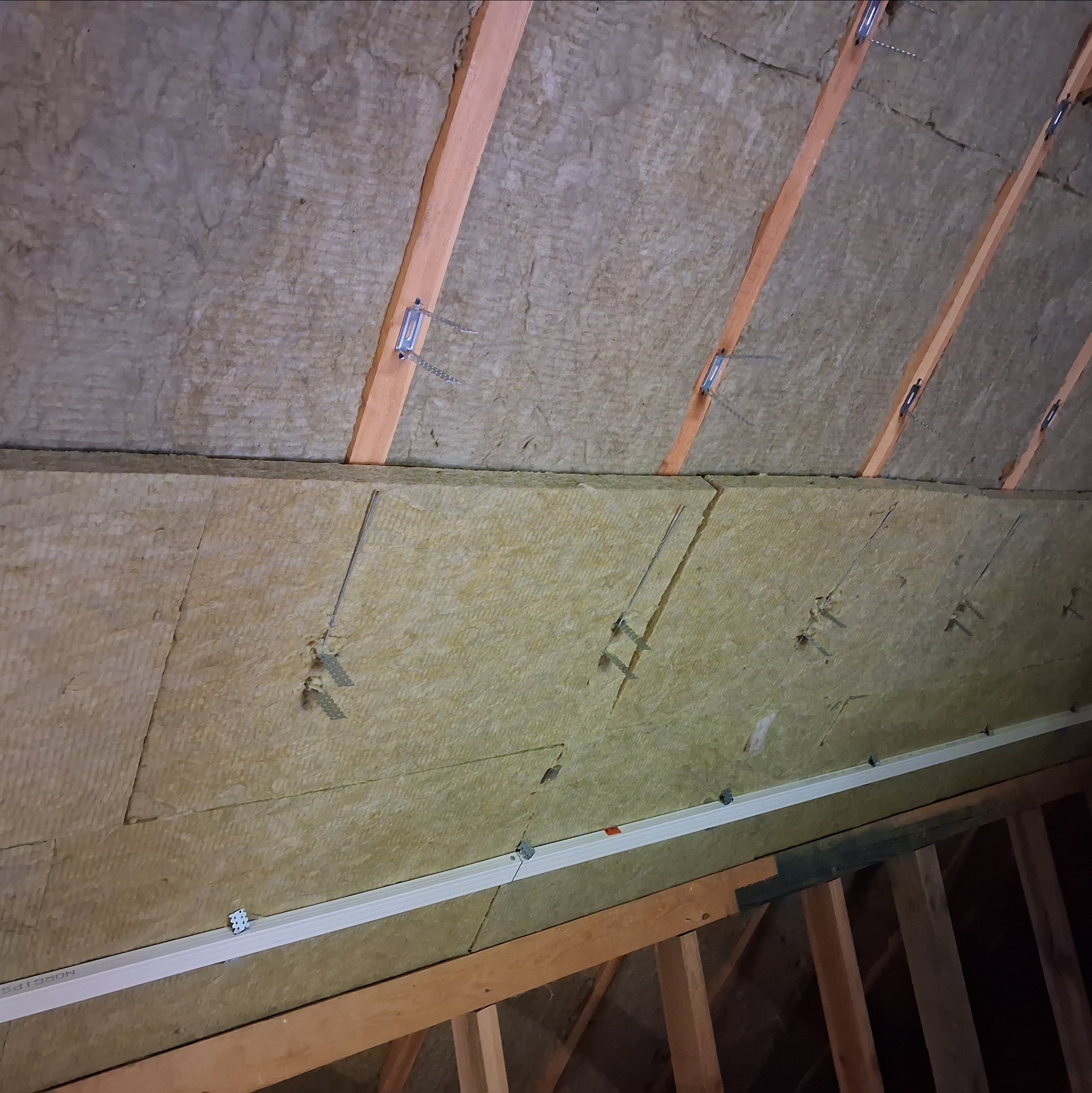 Ocieplenie poddasza wełną mineralną, widoczne drewniane krokwie i metalowe mocowania. Izolacja termiczna w trakcie montażu, widok z dołu na skośny sufit.