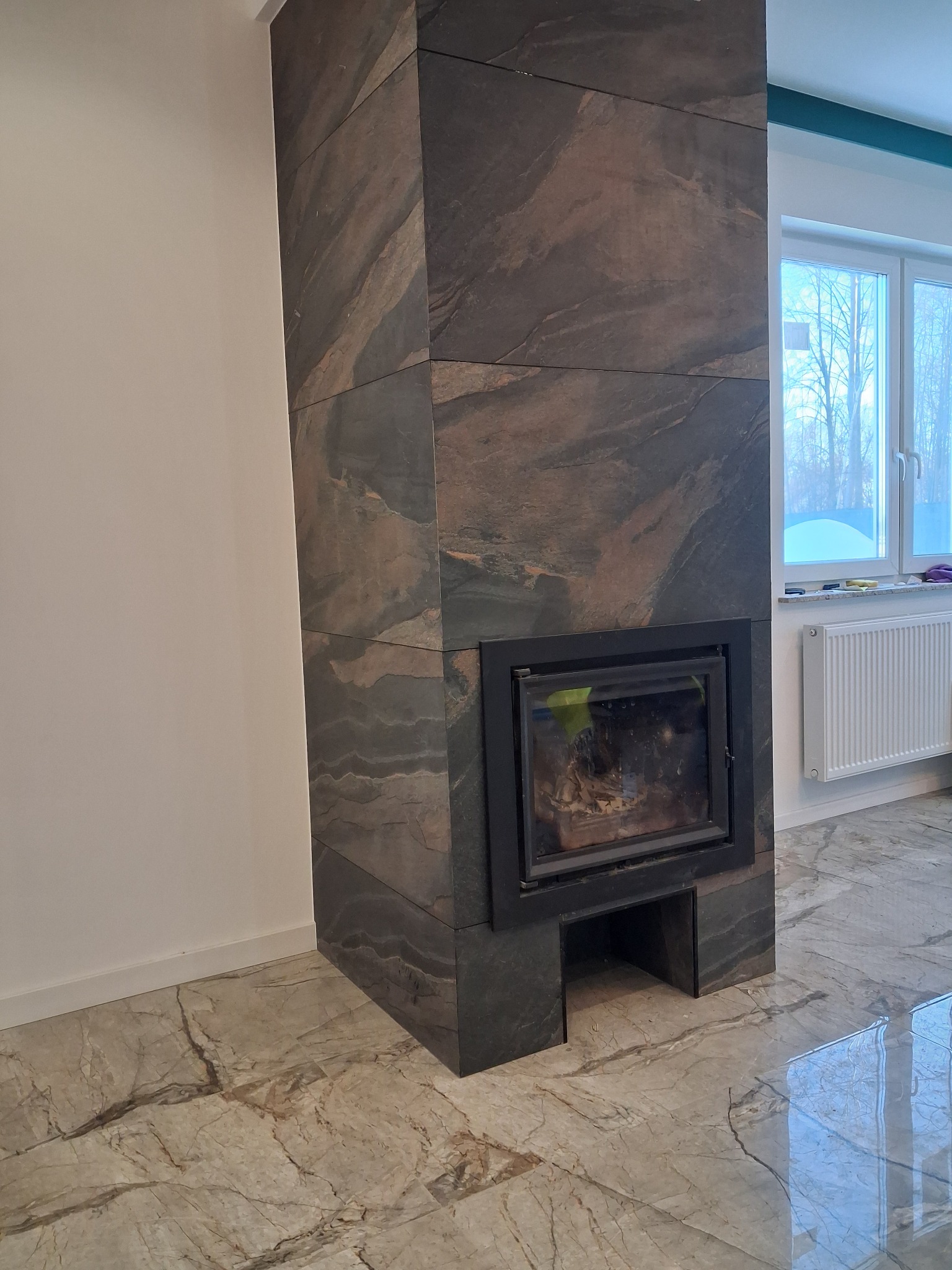 Obudowa kominka z kamienia naturalnego o wyraźnym wzorze żyłkowania, dopasowana do marmurowej podłogi. W tle okno z widokiem na zimowy krajobraz.