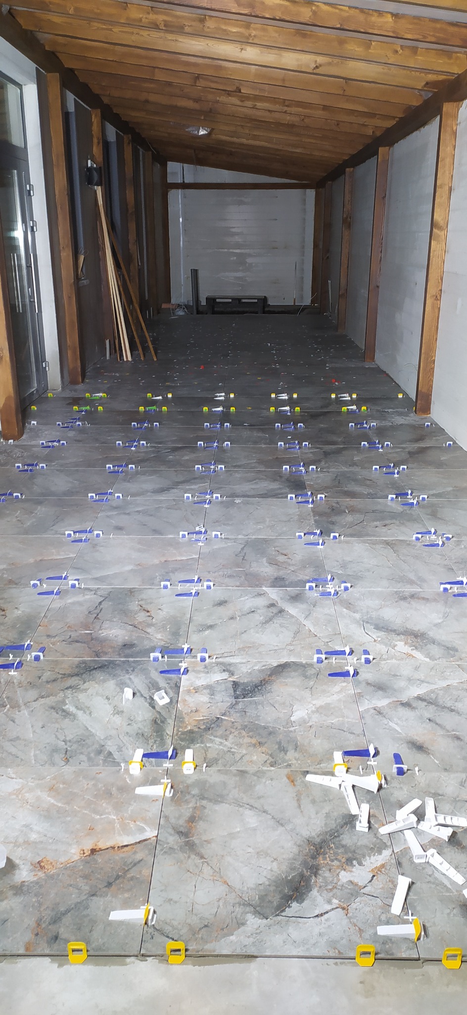 Układanie płytek podłogowych z użyciem klipsów poziomujących w długim pomieszczeniu z drewnianą konstrukcją sufitu. Widok z perspektywy podłogi.