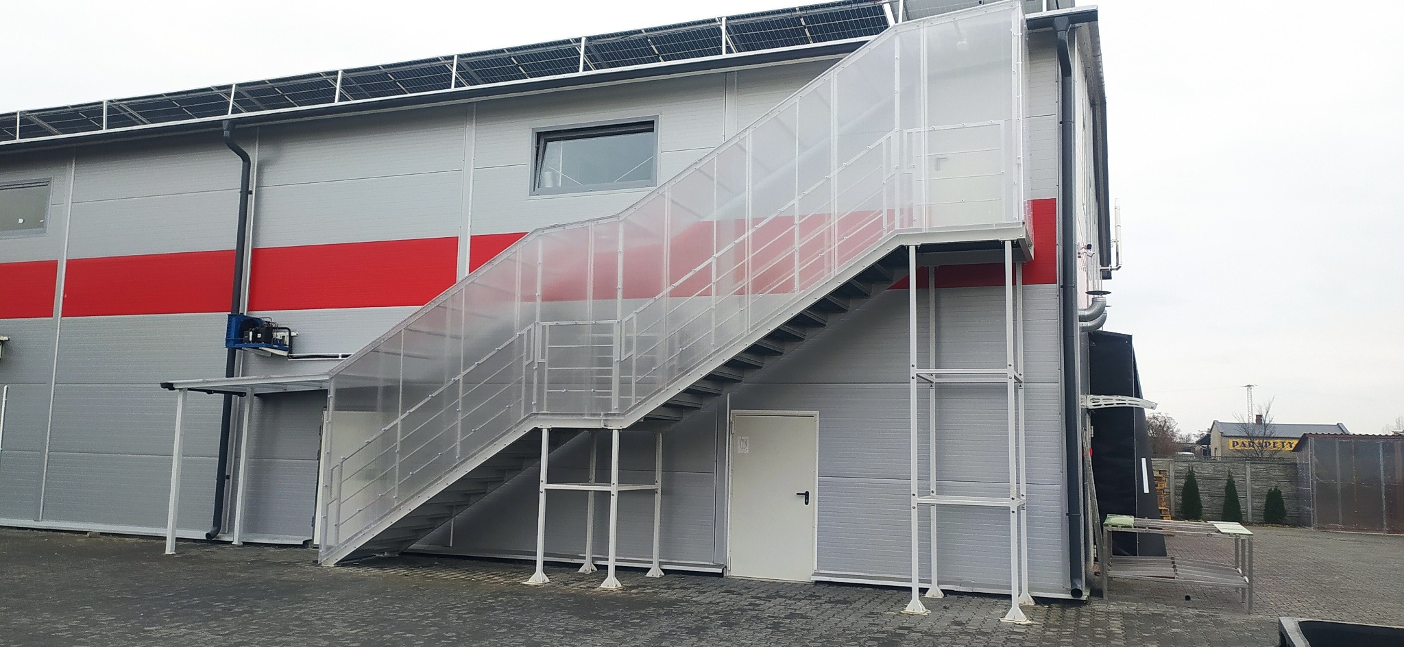 Zewnętrzne schody metalowe z przezroczystą osłoną, prowadzące do budynku z panelami słonecznymi na dachu i czerwoną linią. Widok z dołu, pochmurne niebo.