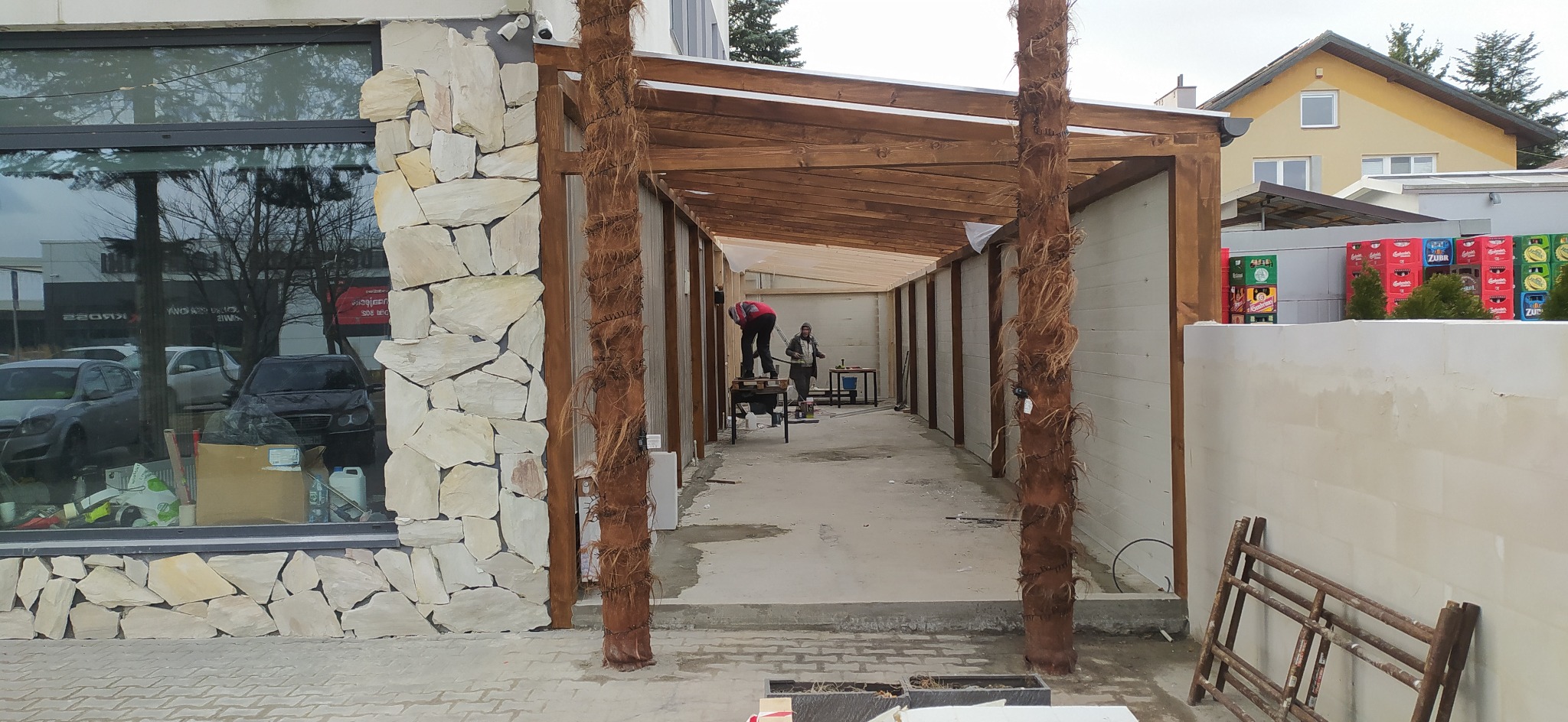 Budowa zadaszenia tarasu z drewnianą konstrukcją i elementami dekoracyjnymi imitującymi palmy. W tle budynek z kamienną elewacją i pracownicy na rusztowaniu.