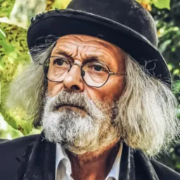 Portret starszego mężczyzny z brodą i kapeluszem, w okularach, na tle zielonych liści. Wyrazisty kadr, skupiony na twarzy i detalu ubioru.