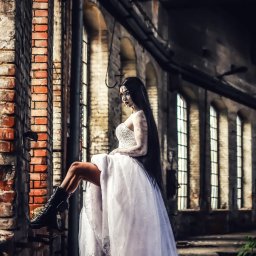 Przemek Paszkiewicz Pasja Obiektywu - Mroczna panna młoda w białej sukni i czarnych butach siedzi na tle ceglanej ściany w opuszczonym budynku z rzędami okien. Makijaż i stylizacja tworzą gotycki klimat.