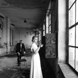 Przemek Paszkiewicz Pasja Obiektywu - Czarno-białe zdjęcie pary w opuszczonym budynku. Kobieta w białej sukni wskazuje palcem na ścianę, mężczyzna w garniturze opiera się o krzesło w tle. Surowy klimat.