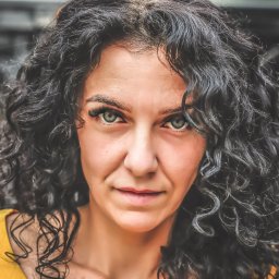 Przemek Paszkiewicz Pasja Obiektywu - Portret kobiety o przenikliwym spojrzeniu, z burzą ciemnych, kręconych włosów i delikatnym makijażu, ubrana w żółtą bluzkę, na rozmytym, zielonym tle.