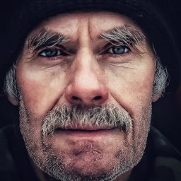 Przemek Paszkiewicz Pasja Obiektywu - Portret starszego mężczyzny z siwymi włosami i wąsami, w czapce, z bliska, z intensywnym spojrzeniem niebieskich oczu, na ciemnym tle. Wyraziste zmarszczki i detale skóry.