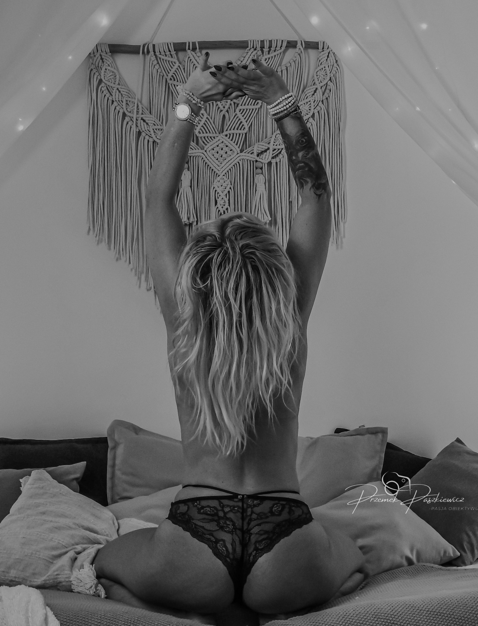 Kobieca sesja w bieliźnie: Modelka siedzi na łóżku tyłem, ręce uniesione, ozdobione bransoletkami i zegarkiem. W tle makrama i delikatne oświetlenie.