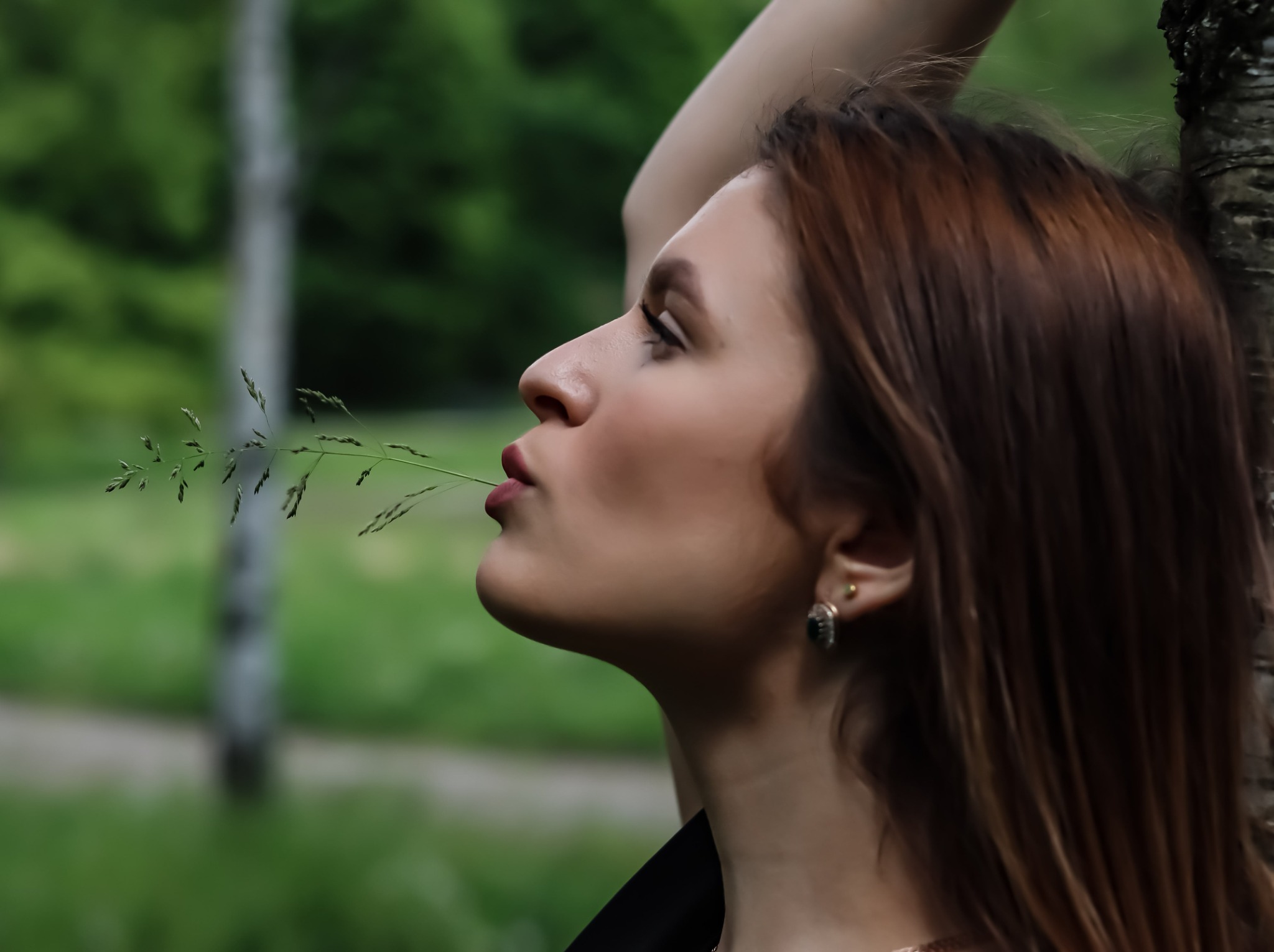 Profil kobiety z źdźbłem trawy w ustach, opierającej się o drzewo na tle zieleni. Ujęcie z dołu, naturalne światło, widoczne kolczyki i delikatny makijaż.