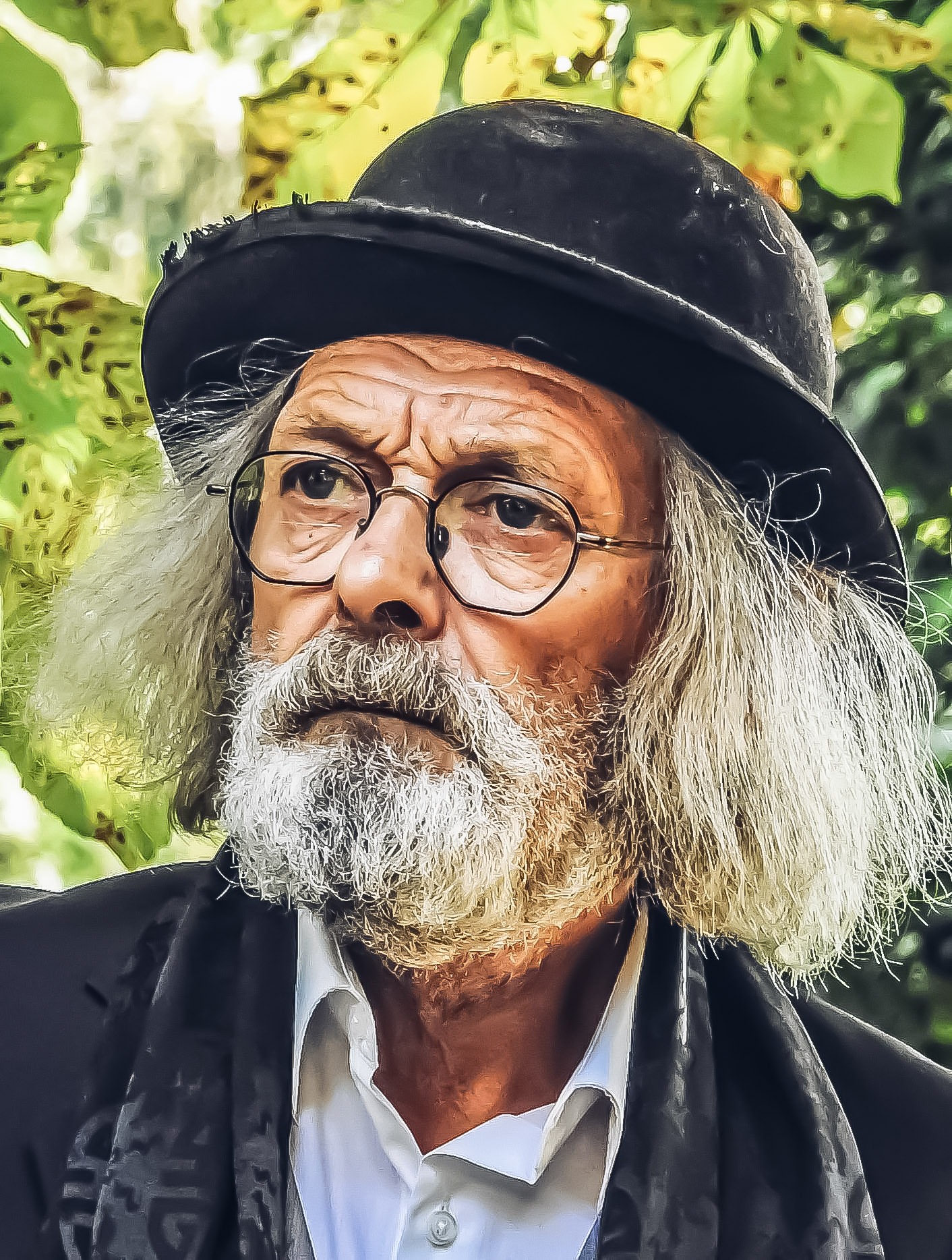 Portret starszego mężczyzny z brodą i kapeluszem, w okularach, na tle zielonych liści. Wyrazisty kadr, skupiony na twarzy i detalu ubioru.