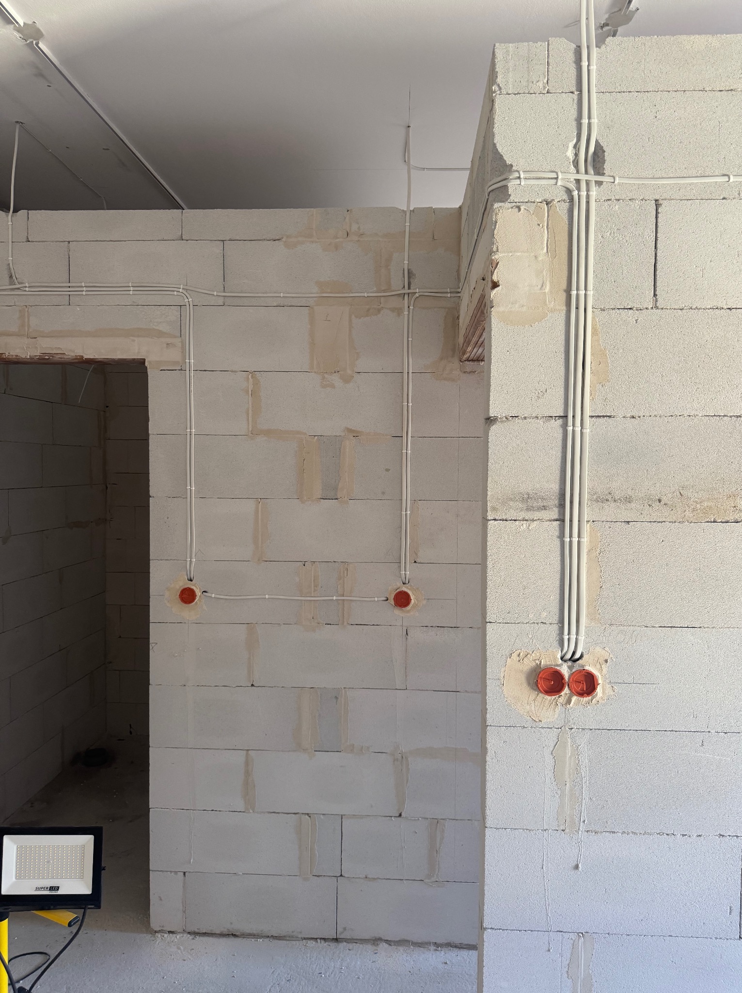 Wewnętrzne instalacje elektryczne w surowym budynku z bloków, widoczne kable i puszki elektryczne, oświetlenie budowlane w rogu. Etap prac przed wykończeniem.