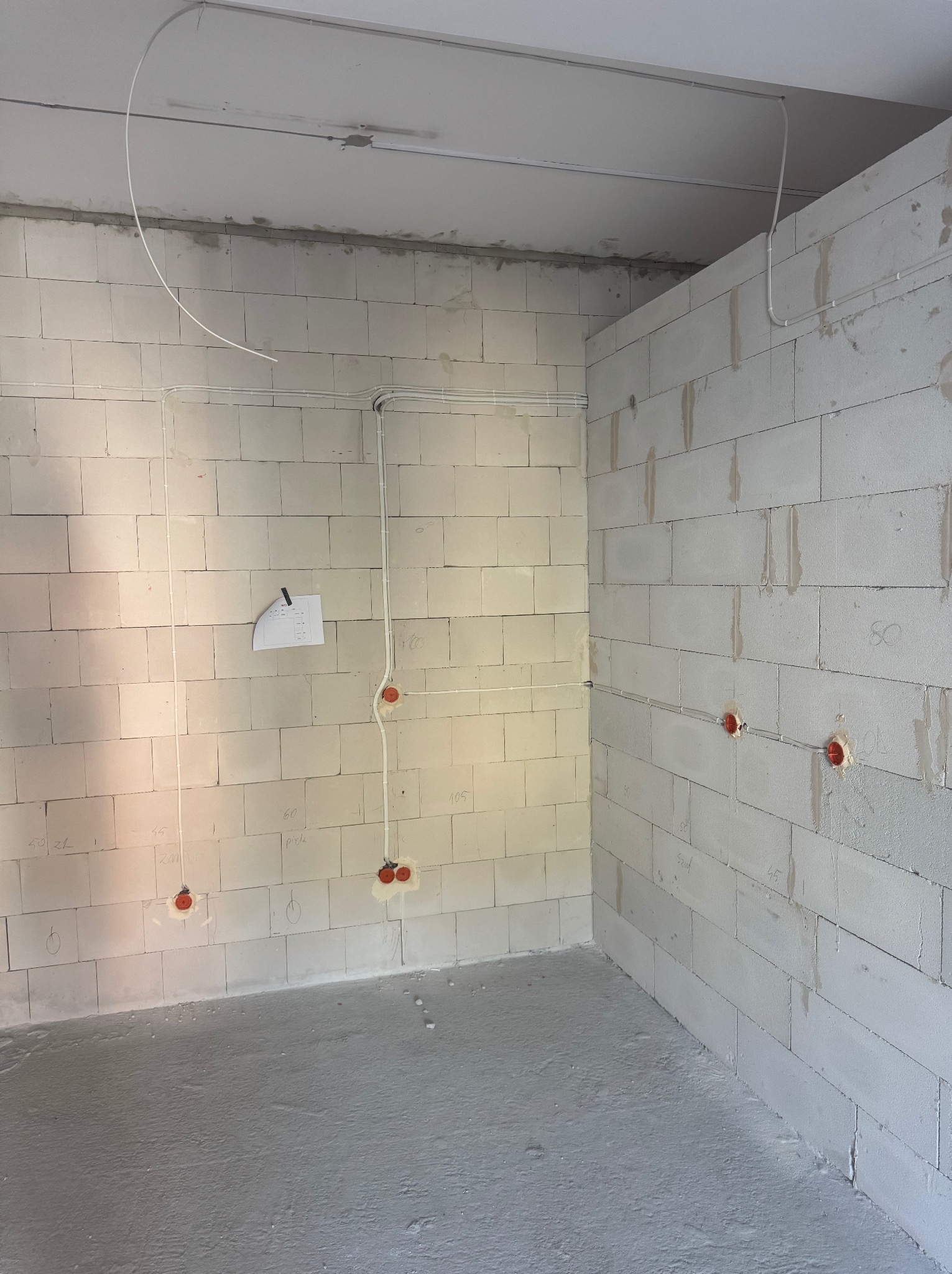 Surowy stan: białe ściany z betonu komórkowego z widoczną, częściowo rozprowadzoną instalacją elektryczną z pomarańczowymi puszkami i białymi kablami.
