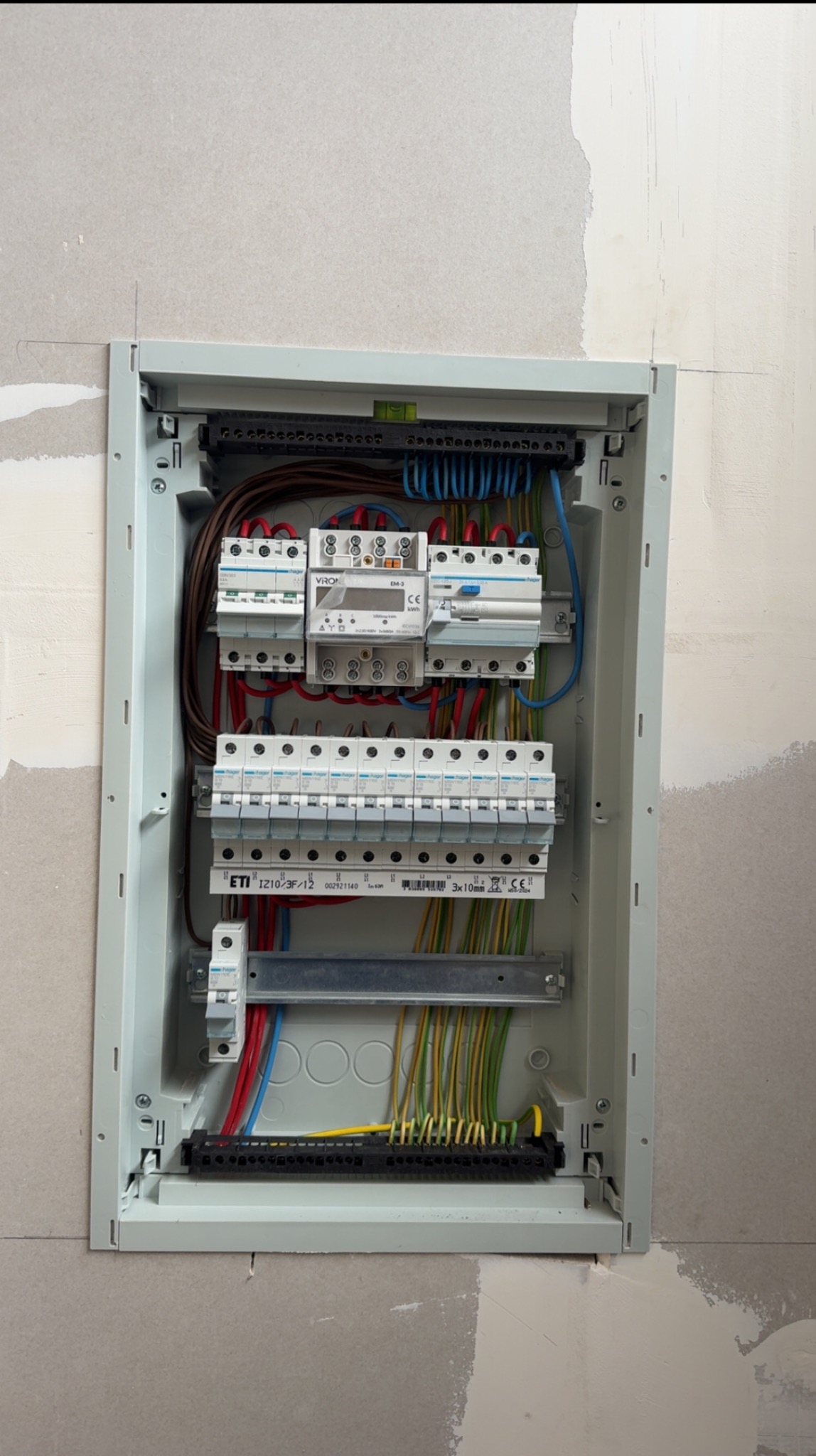 Rozdzielnica elektryczna w szarej obudowie, zamontowana w ścianie, z widocznymi przewodami, bezpiecznikami i licznikiem energii. Instalacja w trakcie prac wykończeniowych.