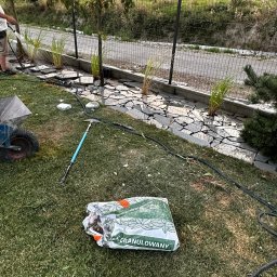 Sekwoja Garden - Zakładanie ogrodu: sadzenie roślin wzdłuż ogrodzenia, nawadnianie, rozsypana ziemia granulowana, narzędzia ogrodnicze, widok na wzgórza w tle.