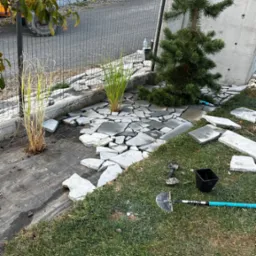 Układanie nieregularnych płyt kamiennych w ogrodzie, widoczne narzędzia ogrodnicze i sadzonki traw ozdobnych, w tle siatka ogrodzeniowa i betonowy mur.
