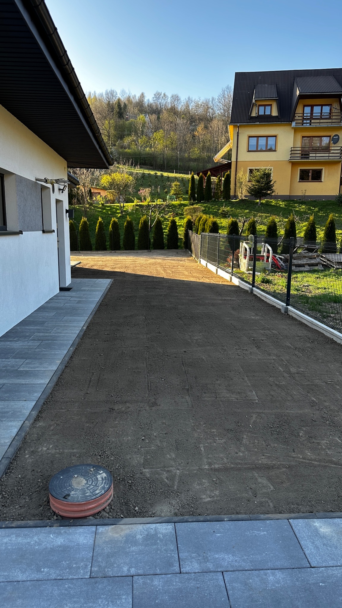 Przygotowany teren pod trawnik z rolki, widok z boku domu, w tle budynek i drzewa. Teren wyrównany, widoczne ślady po pracy glebogryzarki. Słoneczny dzień.