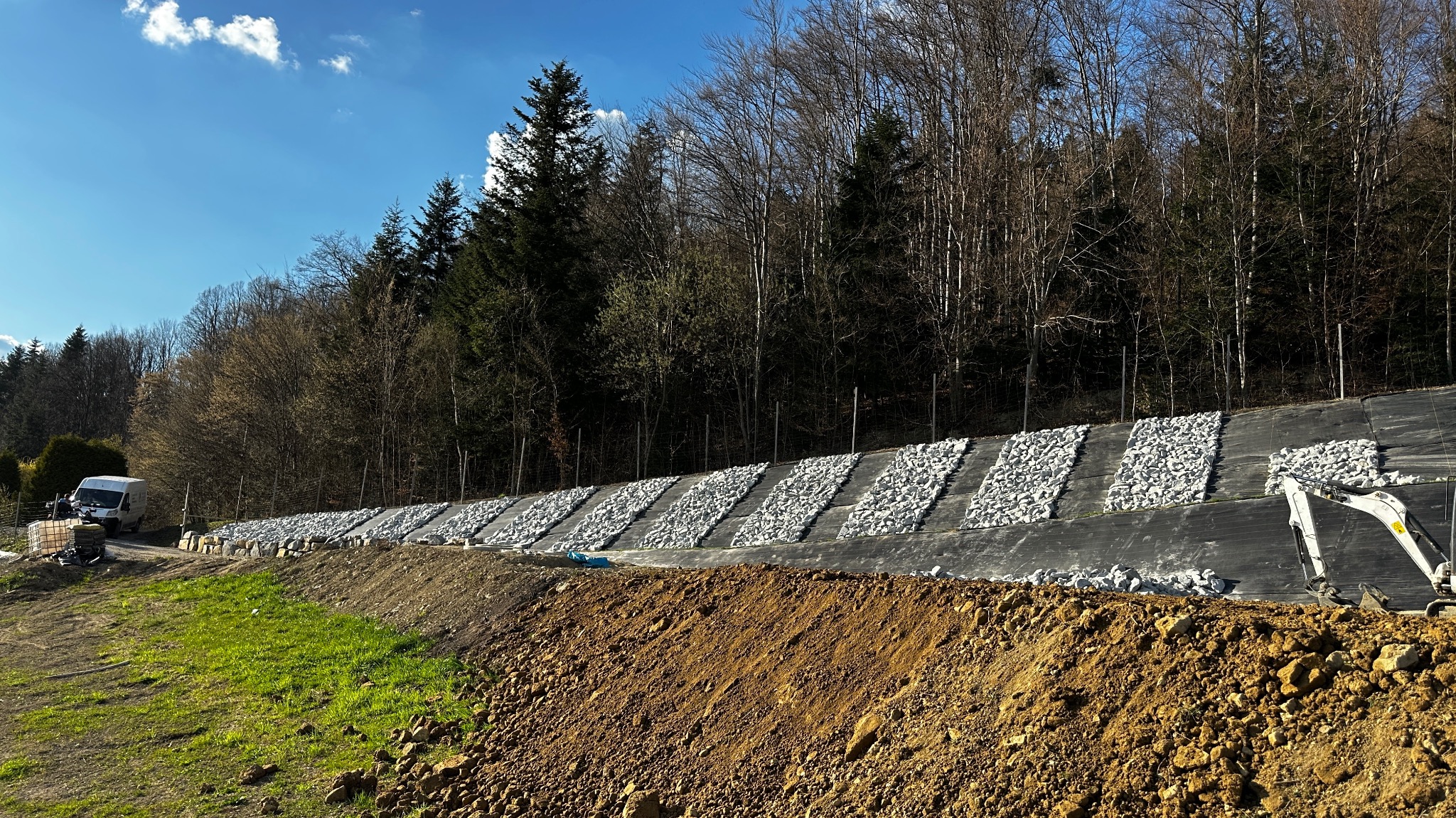 Skarpa zabezpieczona geowłókniną i pasami z białego kamienia, widoczna koparka i bus. W tle drzewa i niebo. Ujęcie z dołu, podkreślające nachylenie terenu.