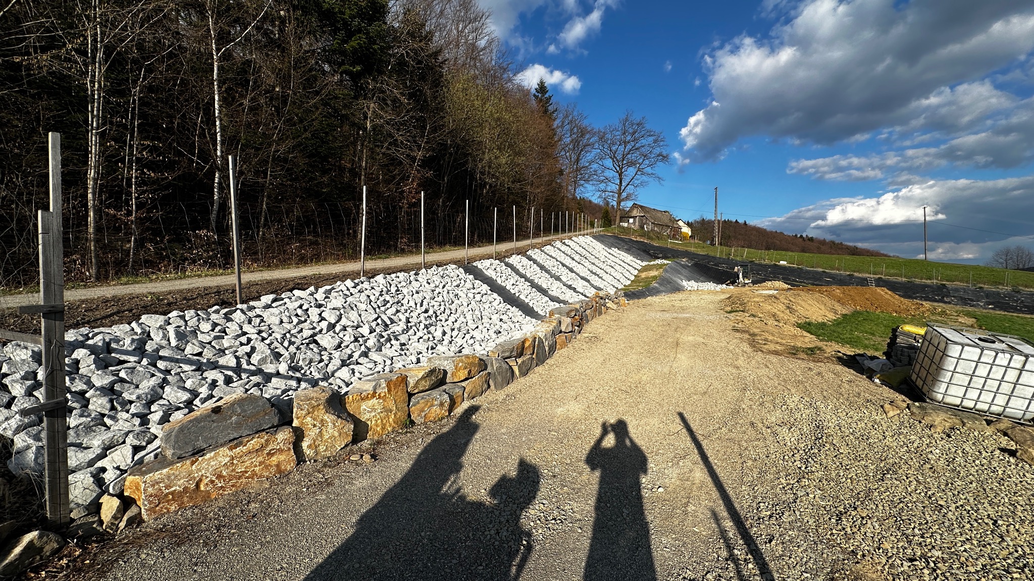 Wzmocnienie skarpy kamieniami z jasnego kruszywa, zabezpieczone murem z kamieni. W tle widok na wzgórze i drzewa. Cień dwóch osób w dolnej części kadru.