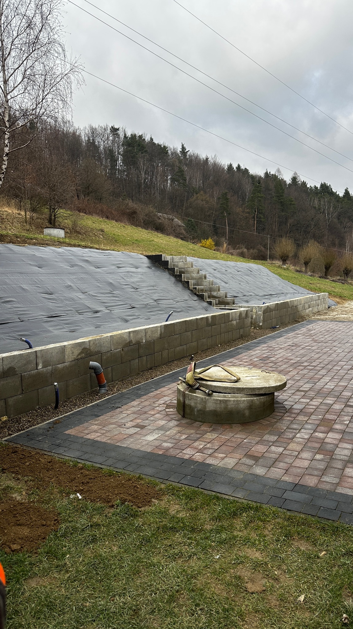 Przygotowany teren pod trawnik z rolki, widoczne schody i studzienka. Częściowo ułożona kostka brukowa. Na górze skarpy czarna mata, zabezpieczenie przed osuwaniem.
