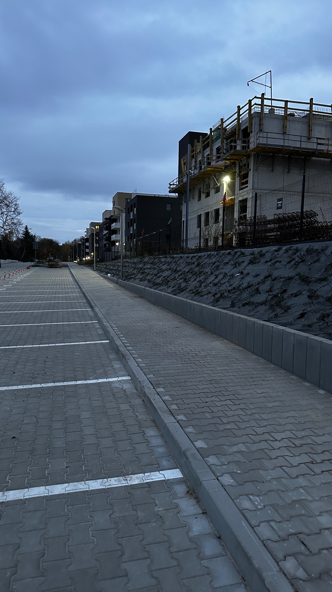 Nowo wybrukowany parking z kostki, białe linie wyznaczające miejsca postojowe. W tle budynek w trakcie budowy, zabezpieczone zbocze. Pochmurne niebo.