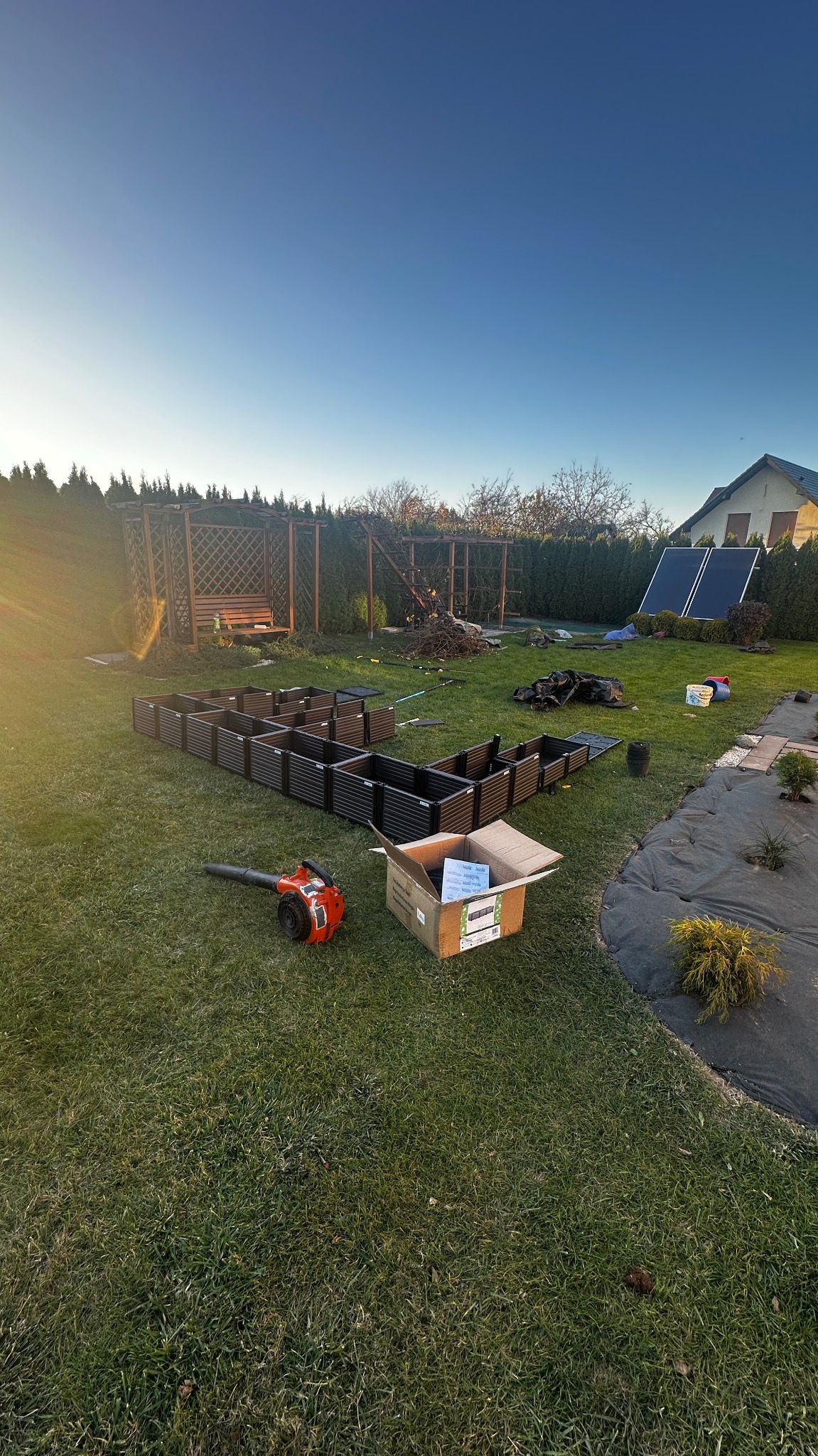 Montaż podwyższonych grządek w ogrodzie, widoczne narzędzia i elementy konstrukcyjne. W tle altana i dom. Poranne słońce oświetla zieloną trawę.