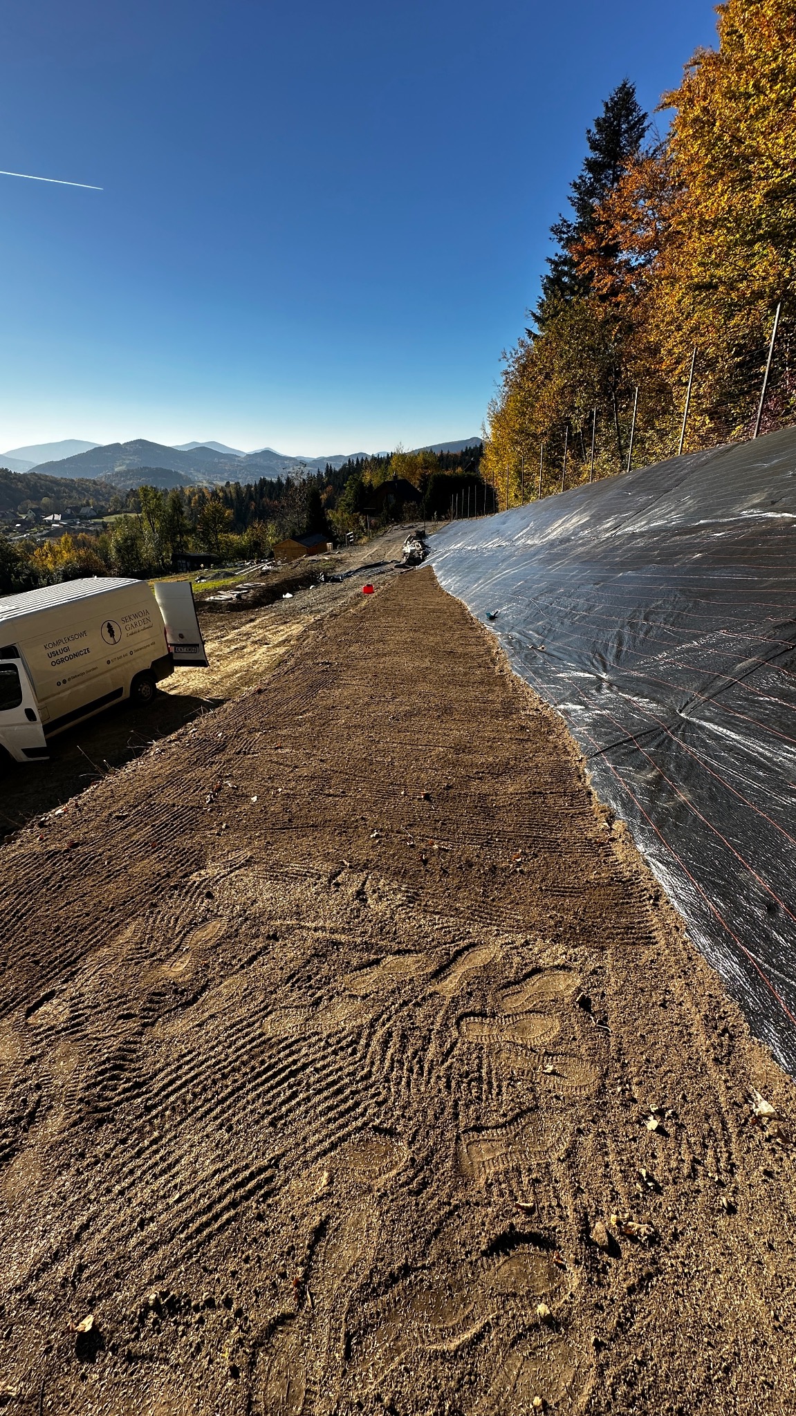 Przygotowanie terenu pod trawnik z rolki na zboczu. Widoczne ślady opon i stóp na ziemi, po prawej stronie folia. W tle las i zabudowania.