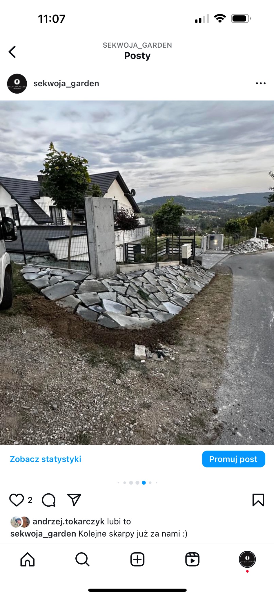 Skarpa z kamienia naturalnego ułożona wzdłuż drogi, z betonowym słupkiem. W tle dom z ogrodem i widok na góry. Efektowna aranżacja przestrzeni.