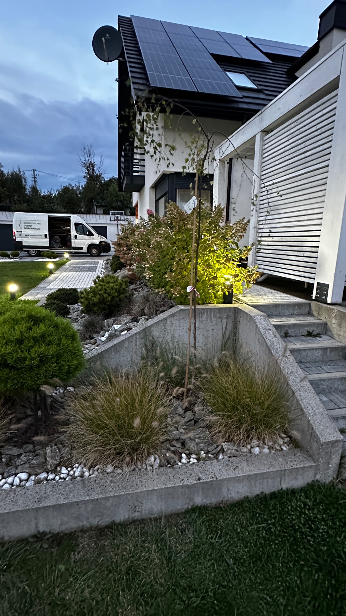Ogród z niską roślinnością ozdobną, kamieniami i betonowym obrzeżem, z oświetlonymi ścieżkami i nowoczesnym domem z panelami słonecznymi w tle.