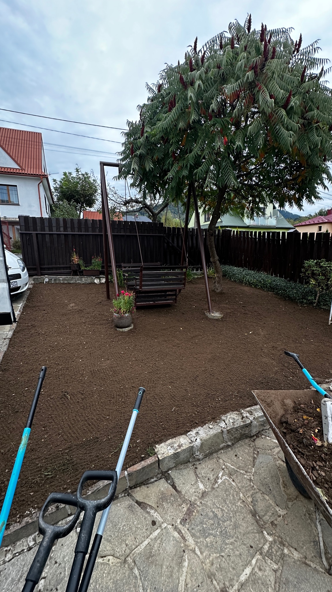 Przygotowany teren pod trawnik z rolki, widoczna ziemia, huśtawka ogrodowa, drzewo i narzędzia ogrodnicze. W tle drewniane ogrodzenie i dom.