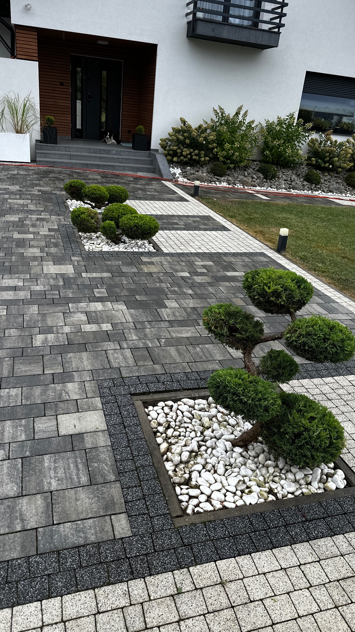 Nowoczesny chodnik z szarej kostki brukowej, urozmaicony rabatami z białych kamieni i formowanymi krzewami. W tle widoczny budynek z elementami drewnianymi.