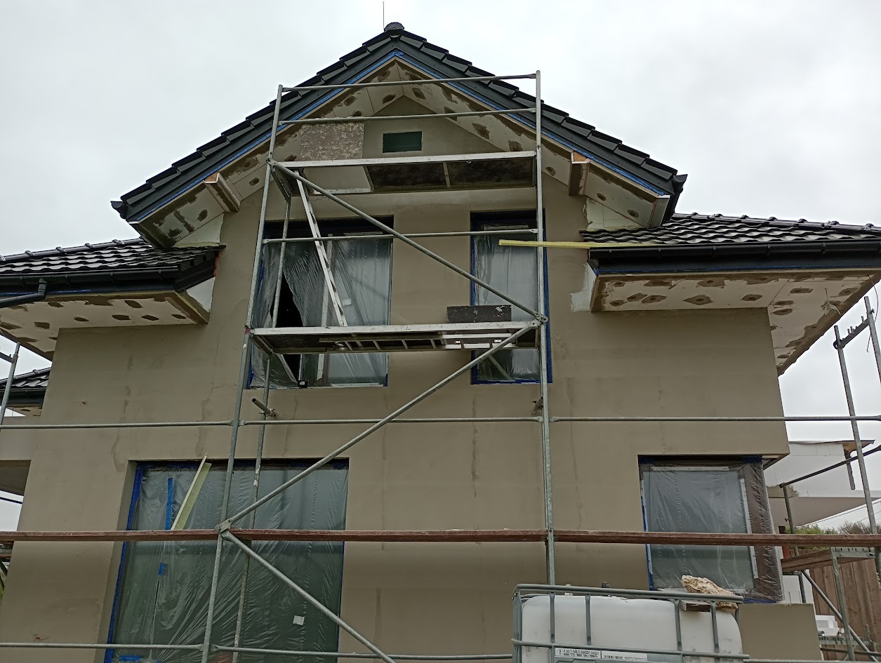 Dom w trakcie ocieplania, widoczne rusztowania i zabezpieczone okna folią. Na elewacji widoczne fragmenty ocieplenia i szpachlowania. Pochmurne niebo w tle.