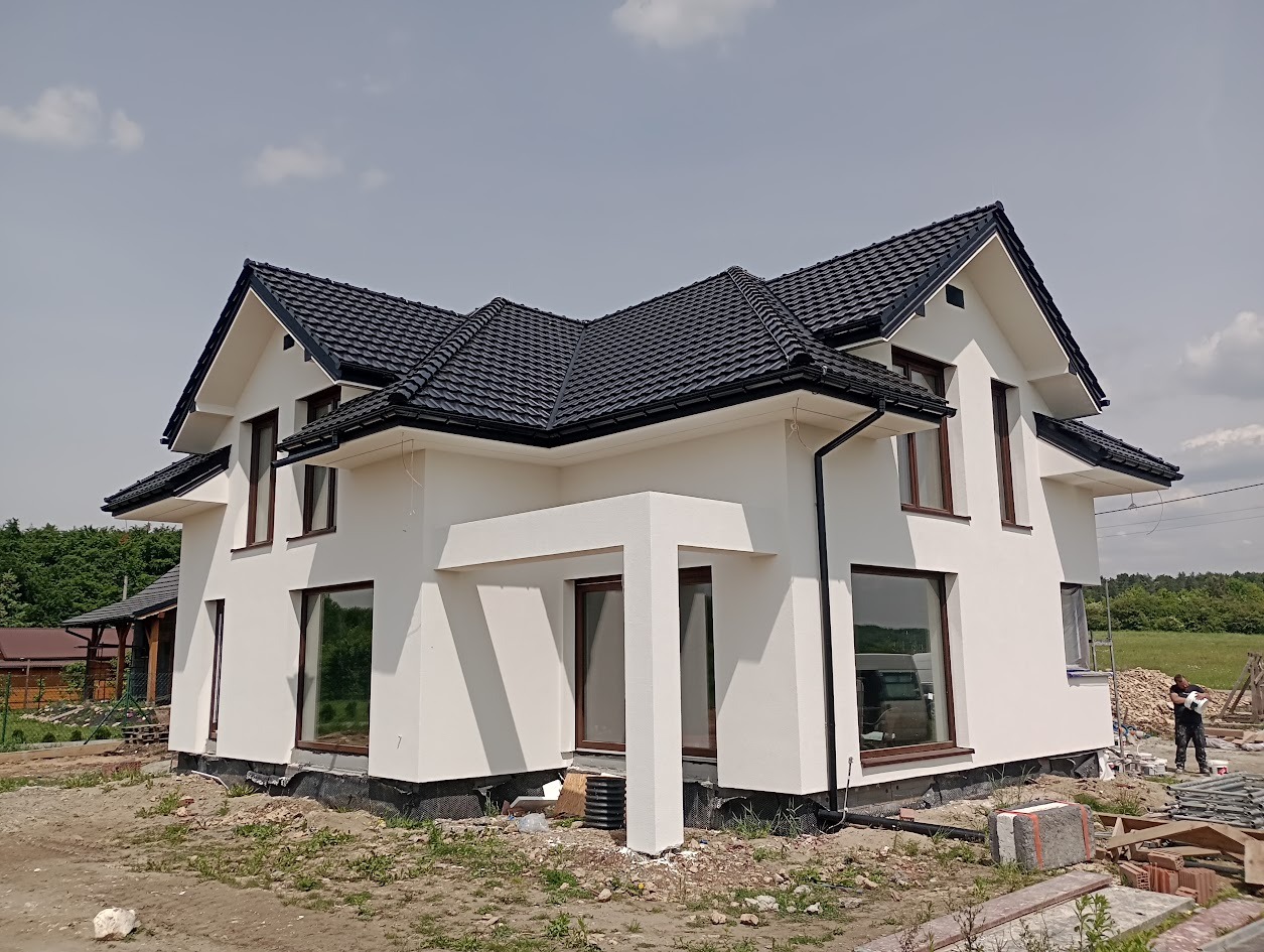 Nowoczesny dom jednorodzinny z białą elewacją i ciemną dachówką, widoczny z boku, z elementami wykończeniowymi w kolorze brązowym.