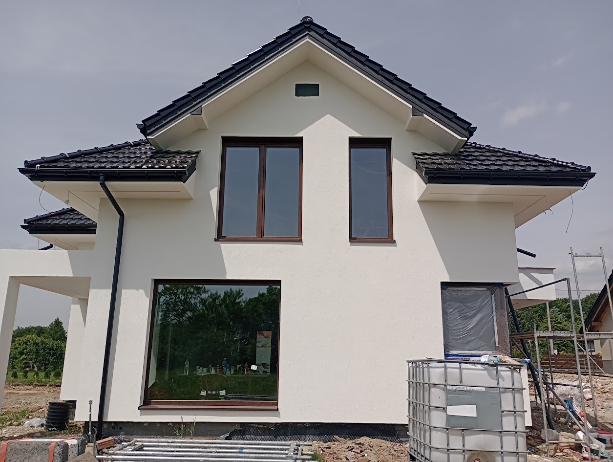 Wykończona elewacja domu jednorodzinnego z ciemną dachówką i brązowymi oknami. Na dole widoczne materiały budowlane i zbiornik na wodę. Część okna zabezpieczona folią.