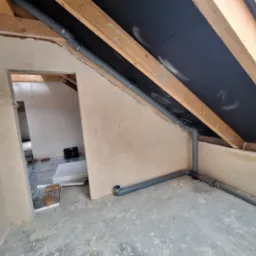 Szare rury kanalizacyjne zamontowane w poddaszu, widoczne drewniane krokwie i czarna membrana dachowa. W tle pomieszczenie z przygotowanym ogrzewaniem podłogowym.