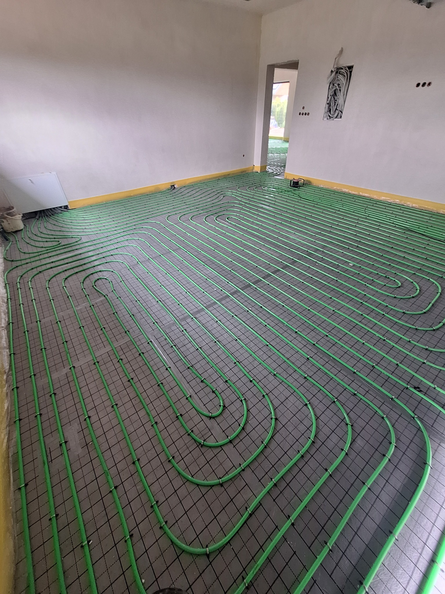 Zielone rury ogrzewania podłogowego ułożone na czarnej siatce w jasnym pomieszczeniu z żółtą taśmą przy ścianach. Widoczny fragment instalacji elektrycznej.