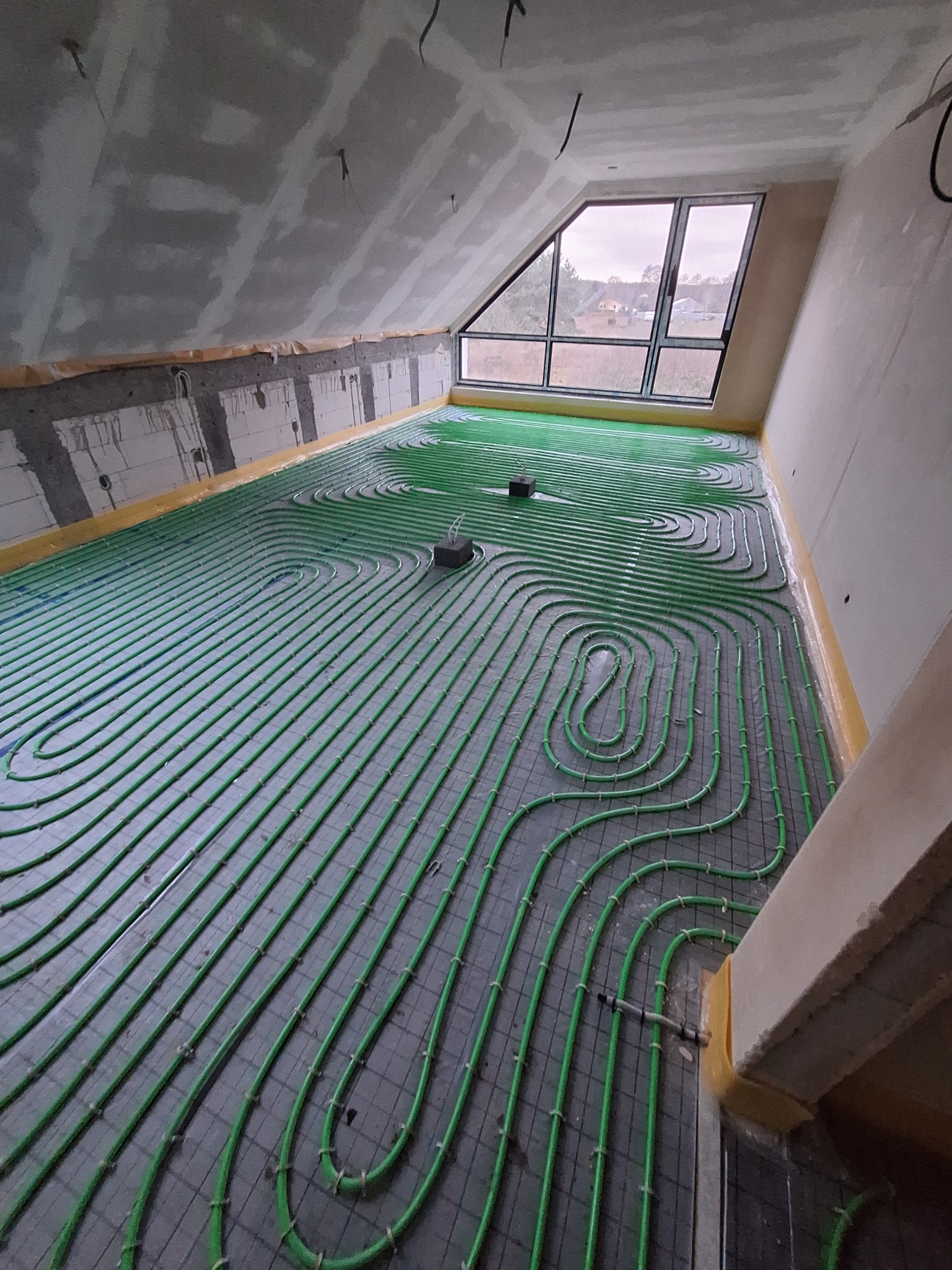 Zielone rury ogrzewania podłogowego ułożone w jodełkę na szarej macie w pokoju na poddaszu z oknem. Widoczne elementy mocujące i żółta taśma wzdłuż ścian.