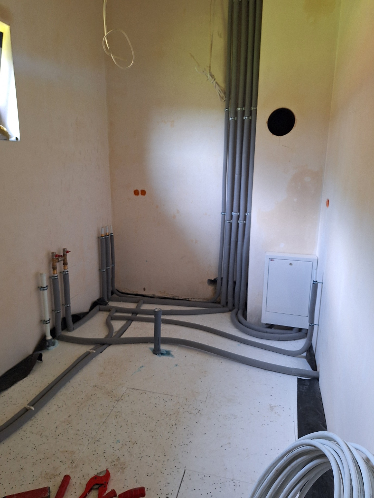 Szare rury instalacji CO ułożone na podłodze i przy ścianach w pomieszczeniu w trakcie budowy. Widoczne narzędzia hydrauliczne.
