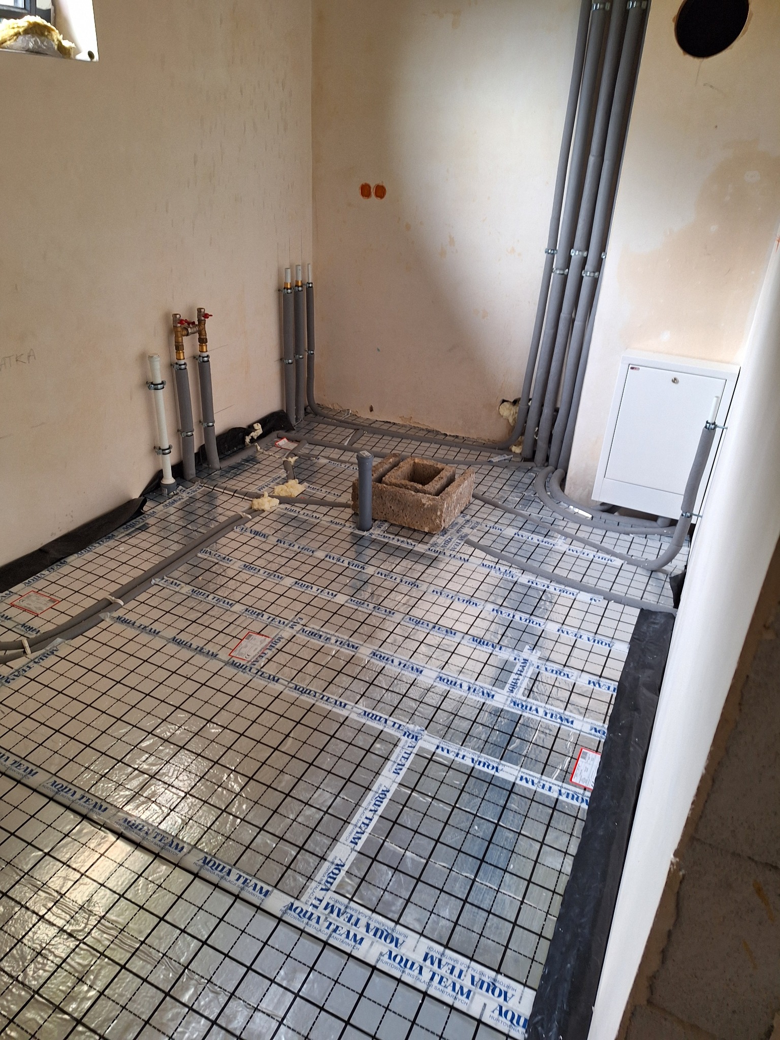 Układanie ogrzewania podłogowego: rury grzewcze na folii z rastrem w surowym wnętrzu. Widoczne piony instalacji i elementy montażowe.