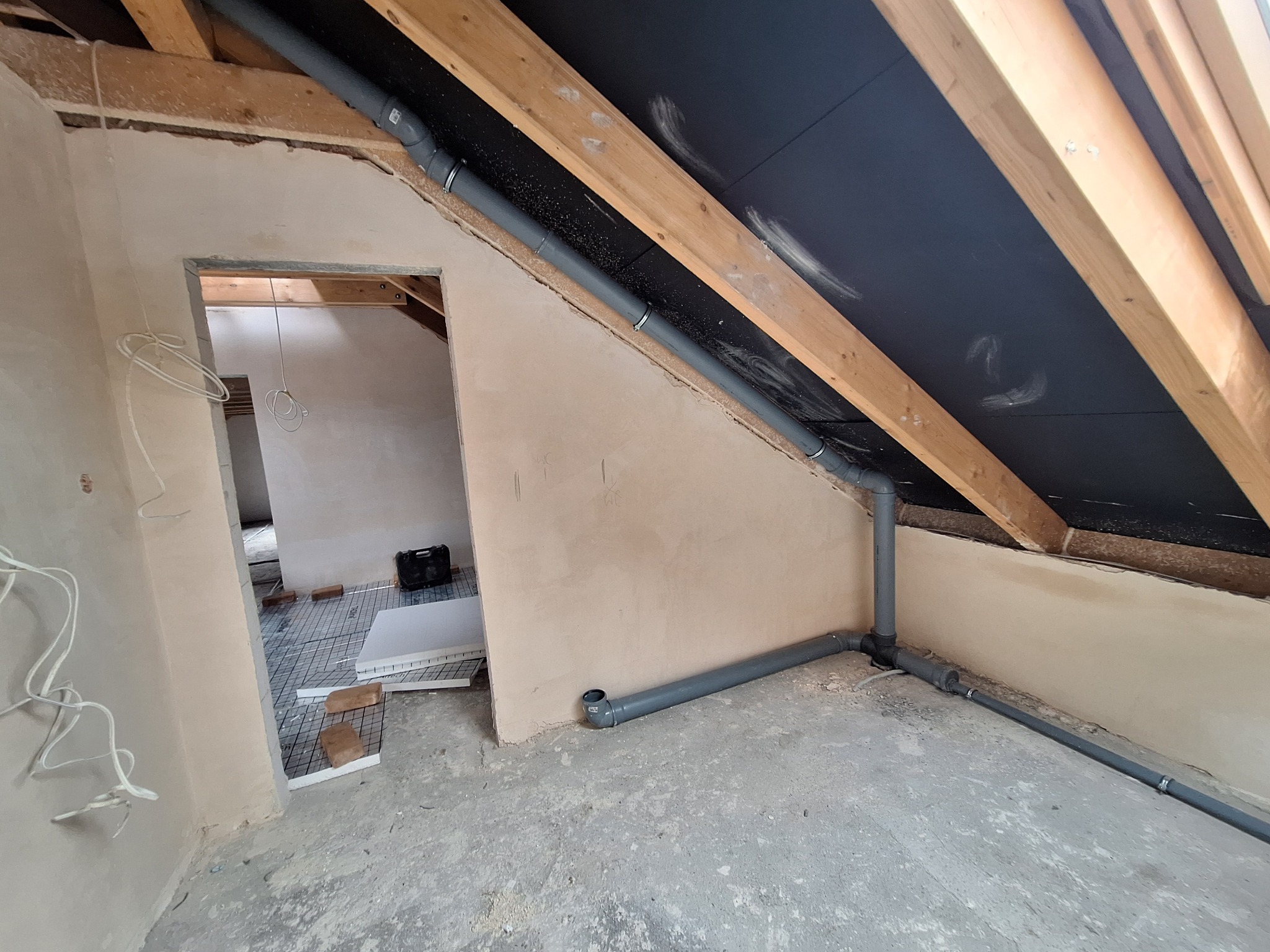 Szare rury kanalizacyjne zamontowane w poddaszu, widoczne drewniane krokwie i czarna membrana dachowa. W tle pomieszczenie z przygotowanym ogrzewaniem podłogowym.