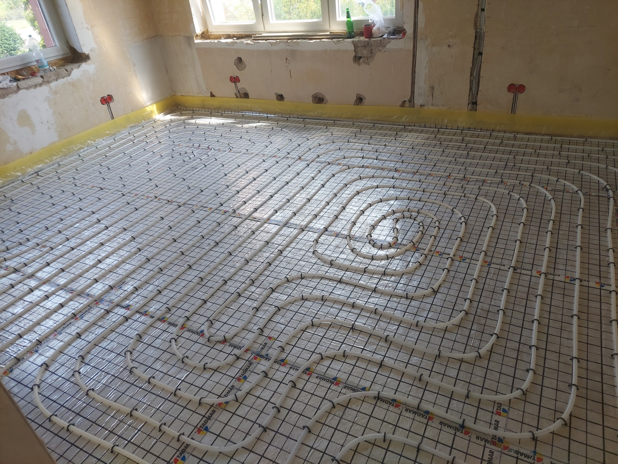 Instalacja ogrzewania podłogowego w trakcie montażu: białe rury ułożone spiralnie na czarnej siatce w surowym wnętrzu z oknem. Żółta taśma przy ścianach.
