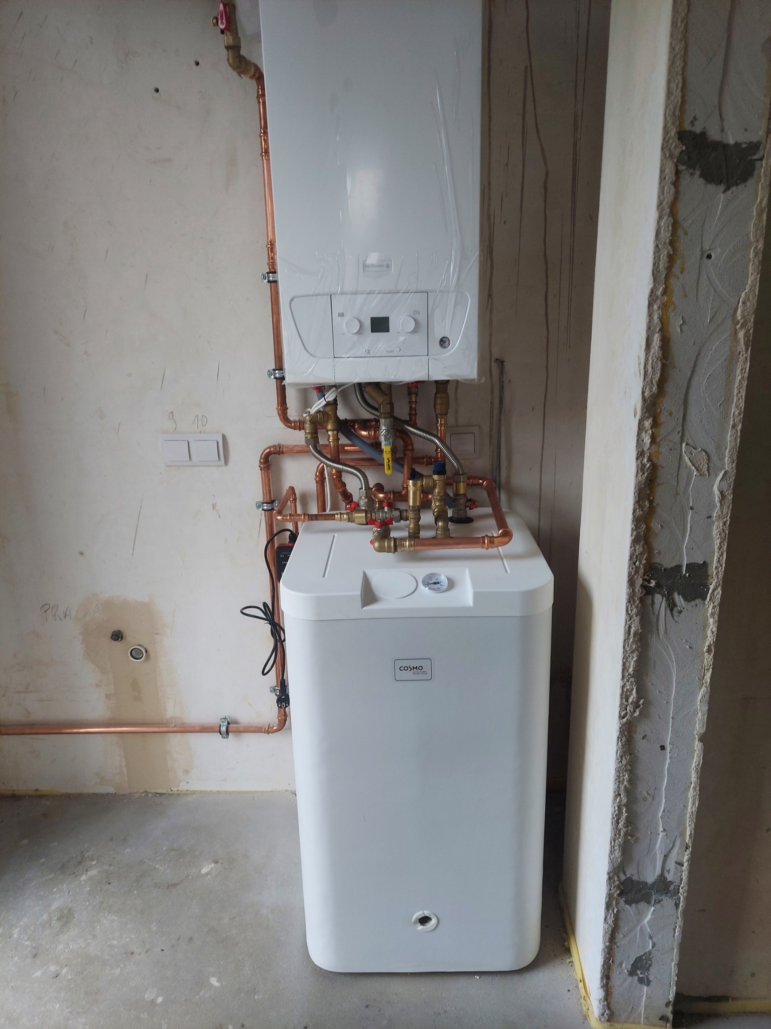 Biały, kondensacyjny piec CO Cosmo z miedzianą instalacją w surowym wnętrzu. Widoczne rury i zawory. Piec częściowo owinięty folią ochronną.