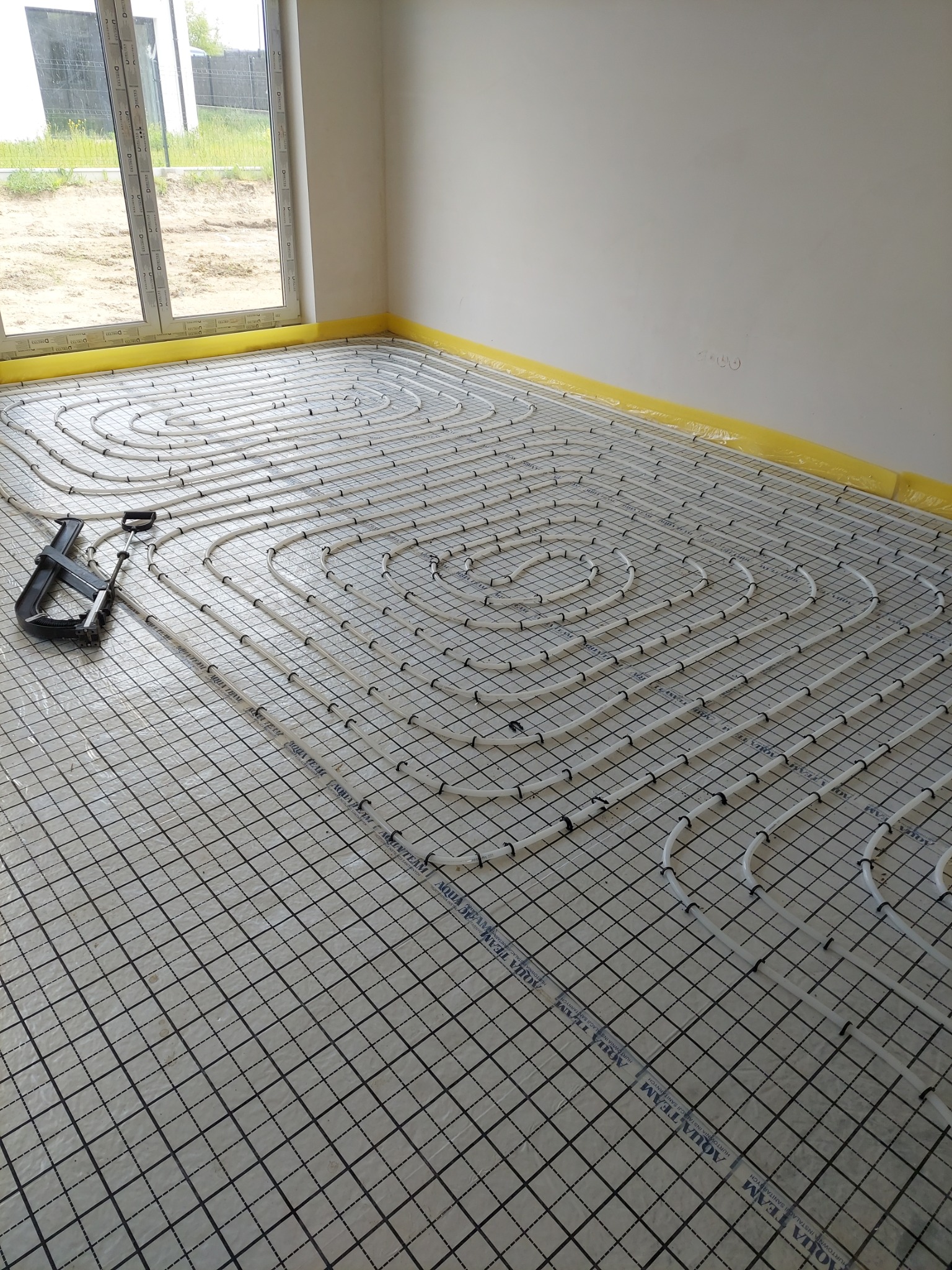 Białe rury ogrzewania podłogowego ułożone spiralnie na czarnej siatce w jasnym pomieszczeniu z oknem. Widoczne narzędzie do mocowania rur.