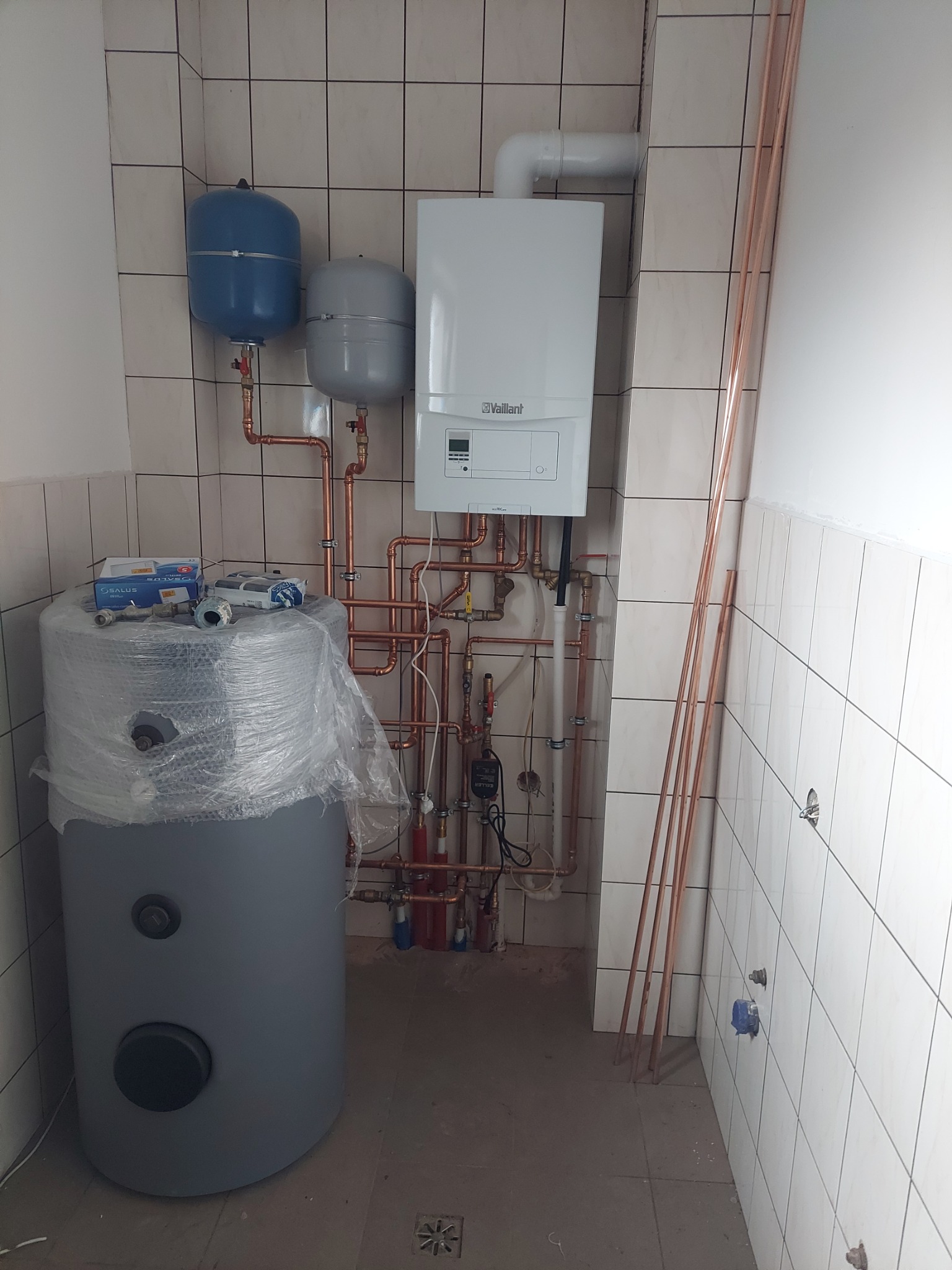 Nowa instalacja CO z piecem Vaillant, zbiornikiem i rurami miedzianymi w pomieszczeniu z kafelkami. Elementy jeszcze częściowo zabezpieczone folią.