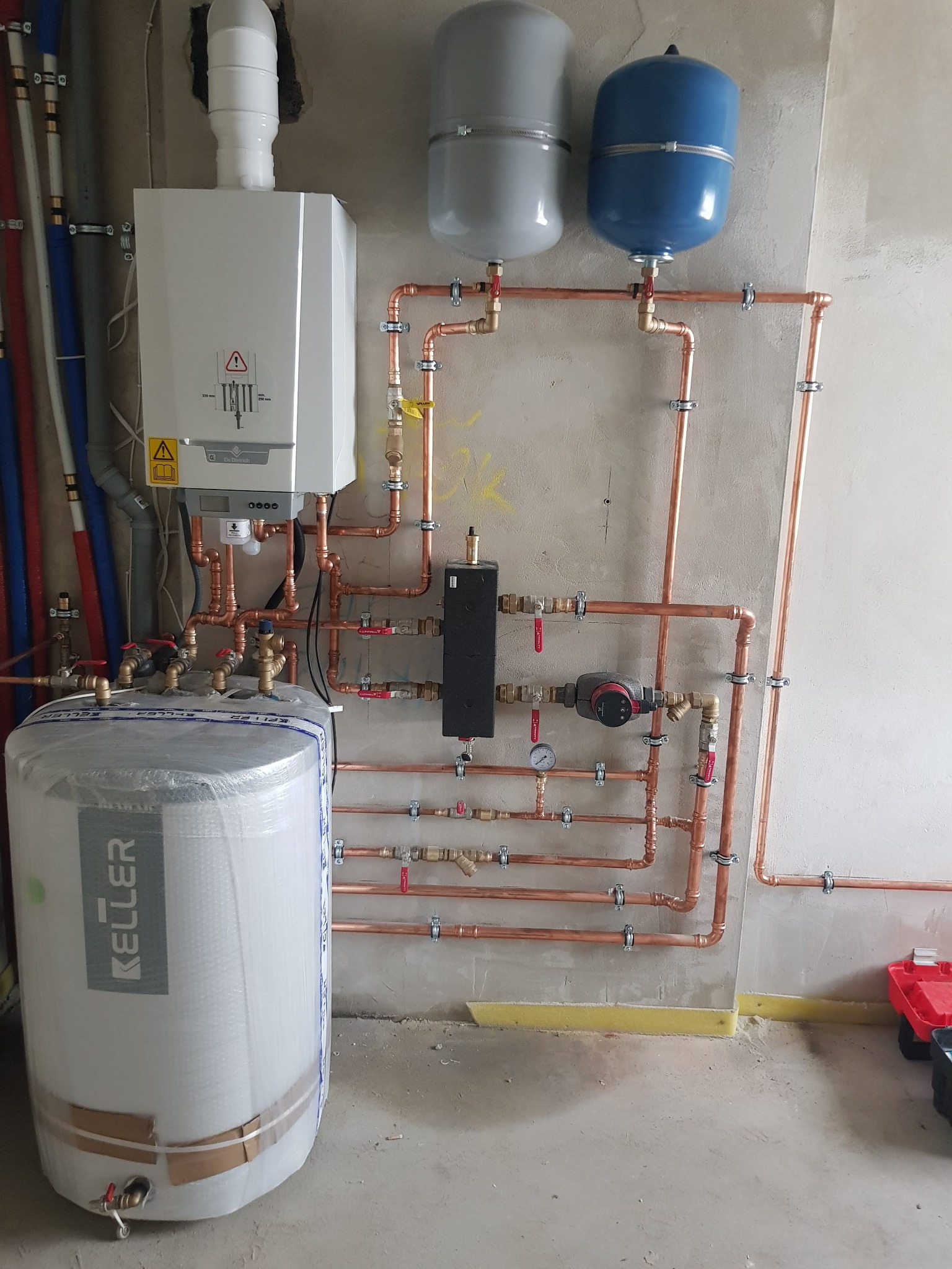 Kompleksowa instalacja grzewcza z piecem De Dietrich, zbiornikiem Keller w folii i miedzianymi rurami na surowej ścianie. Widoczne zawory i manometry.
