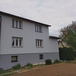 Mixbud - Dwukondygnacyjny dom po renowacji elewacji w odcieniach szarości, z widoczną dolną partią w ciemniejszym kolorze. W tle drzewa i fragmenty innych budynków.